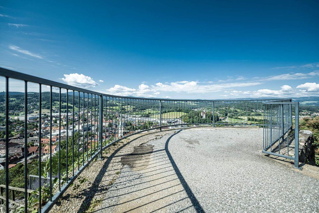 Aussicht von der Burgruine Voitsberg auf das weststeirische Hügelland. Im Vordergrund ein Edelstahl-Geländer zur Absicherung.